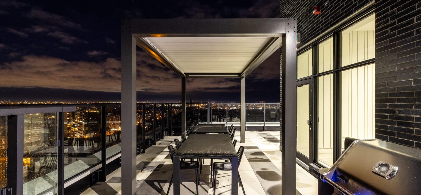 Rooftop terrace at night featuring a modern pergola, dining tables, and a barbecue grill, an exclusive amenity at our luxury apartments in Toronto.